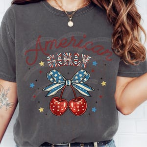 May include: A grey t-shirt with a red, white and blue American flag design. The design features a bow with cherries and the text "American Girl".