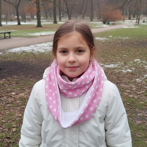 May include: A young person wearing a white jacket and a pink infinity scarf with white polka dots. The scarf is wrapped around the neck. The background shows a park with trees and a walking path.