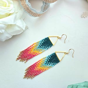 May include: A pair of colorful beaded earrings with a geometric design. The earrings feature a gold triangle top and long fringe in shades of pink, yellow, teal, and blue. The earrings are displayed on a white surface with floral and ribbon accents.