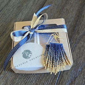May include: A pair of beaded earrings with a gold triangle frame and blue and gold fringe, presented in a gift box tied with a blue and gold ribbon. The box has a Petunia Creations logo.