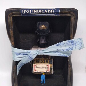 May include: A small wooden box containing a glass bottle with a black cap and a gold emblem. A blue ribbon with white lettering is tied around the bottle. The box has a label that reads "USO INDICADO". A small blue figurine of a person stands in front of the bottle. The bottle has a label that says "Jubilado" and "RECIBIE COPIA".