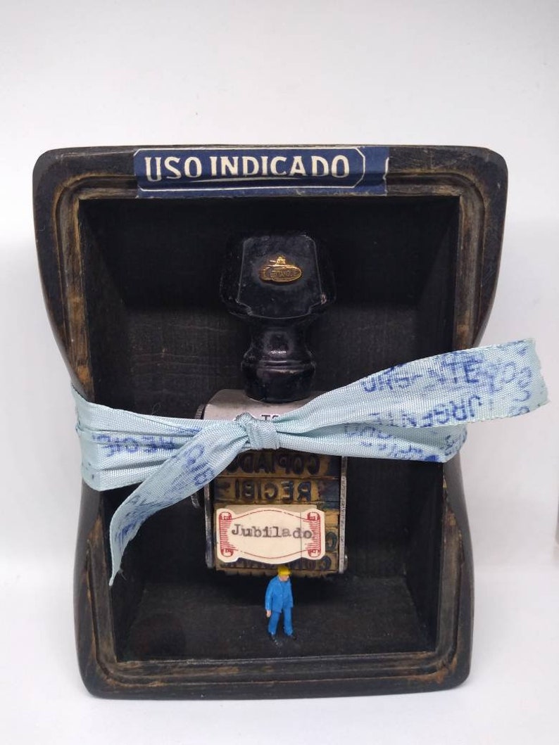 May include: A small wooden box containing a glass bottle with a black cap and a gold emblem. A blue ribbon with white lettering is tied around the bottle. The box has a label that reads "USO INDICADO". A small blue figurine of a person stands in front of the bottle. The bottle has a label that says "Jubilado" and "RECIBIE COPIA".