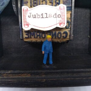 May include: A small, blue figurine of a person standing in front of a vintage wooden stamp with the word "Jubilado" printed on a white label. The stamp has other words printed on it, including "RECIBI" and "COPIADO".