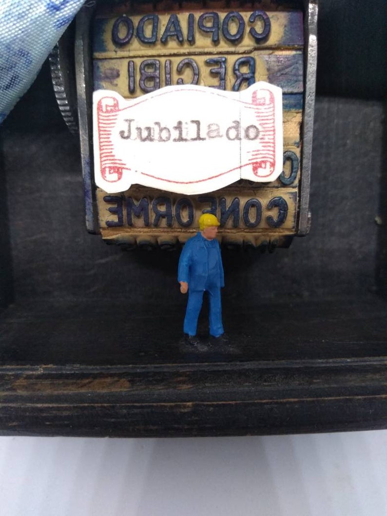 May include: A small, blue figurine of a person standing in front of a vintage wooden stamp with the word "Jubilado" printed on a white label. The stamp has other words printed on it, including "RECIBI" and "COPIADO".