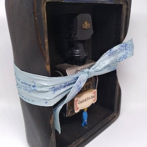 May include: A wooden box with a glass window, containing a small ink bottle, a stamp, and a miniature figure. The box is tied with a blue ribbon and has a label that reads "USO INDICADO".