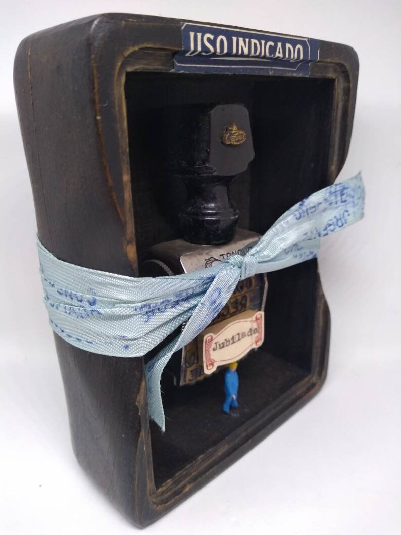 May include: A wooden box with a glass window, containing a small ink bottle, a stamp, and a miniature figure. The box is tied with a blue ribbon and has a label that reads "USO INDICADO".