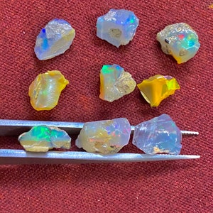 May include: A collection of raw opal gemstones, showcasing a spectrum of colors including blues, greens, yellows, and iridescent flashes. The opals are irregularly shaped and appear to be freshly mined, with rough, natural edges. The gemstones are displayed on a red surface.