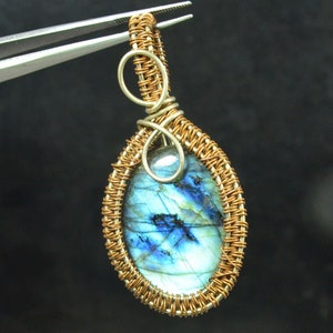 May include: A teardrop-shaped pendant featuring a polished, iridescent blue and grey labradorite gemstone encased in a decorative, intricate copper wire-wrapped frame with a circular bail at the top, held by metal tweezers.