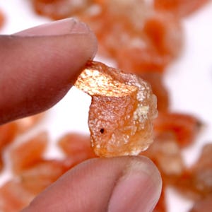 May include: A small, rough-cut, orange-colored gemstone is held between two fingers. The stone has a slightly translucent appearance with a few white inclusions. Other similar stones are visible in the background.