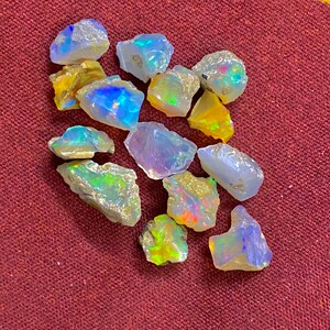 May include: A collection of rough opal gemstones, showcasing a variety of colors including blues, yellows, and greens. The opals have a natural, unpolished appearance, with visible textures and iridescent flashes. The gemstones are scattered on a maroon surface.