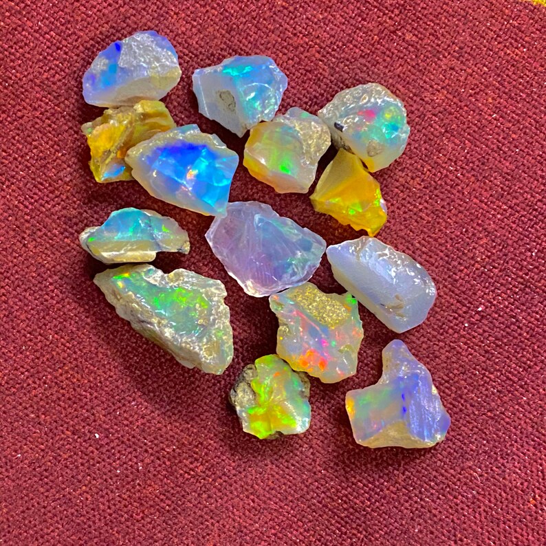 May include: A collection of rough opal gemstones, showcasing a variety of colors including blues, yellows, and greens. The opals have a natural, unpolished appearance, with visible textures and iridescent flashes. The gemstones are scattered on a maroon surface.