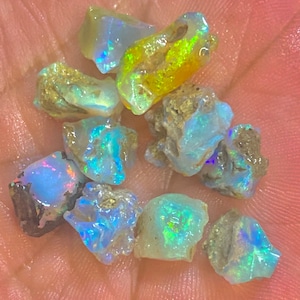 Op de afbeelding: Een verzameling ruwe opaal edelstenen, met een verscheidenheid aan kleuren, waaronder blauw, groen, geel en iriserende tinten. De ruwe stenen hebben een natuurlijke, ongepolijste uitstraling, met een mix van ondoorzichtige en doorschijnende secties.