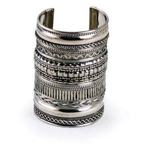 Peut inclure: Un large bracelet manchette argenté avec des motifs texturés complexes. Le bracelet présente divers motifs, notamment des éléments perlés, tressés et géométriques. Le bracelet est ouvert en haut et en bas. Le bijou est présenté sur fond blanc. Circonférence du poignet: environ 16 cm.