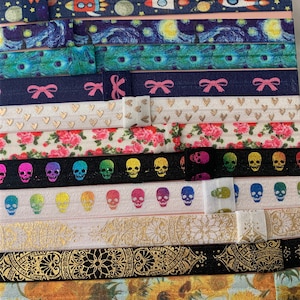 May include: A collection of colorful and patterned hair ties. The hair ties feature designs such as a rocket ship, a starry night sky, hearts, flowers, skulls, a rainbow, a mandala, and sunflowers.