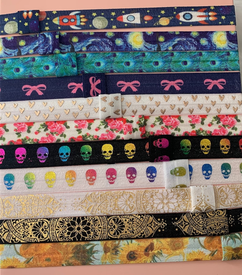 May include: A collection of colorful and patterned hair ties. The hair ties feature designs such as a rocket ship, a starry night sky, hearts, flowers, skulls, a rainbow, a mandala, and sunflowers.