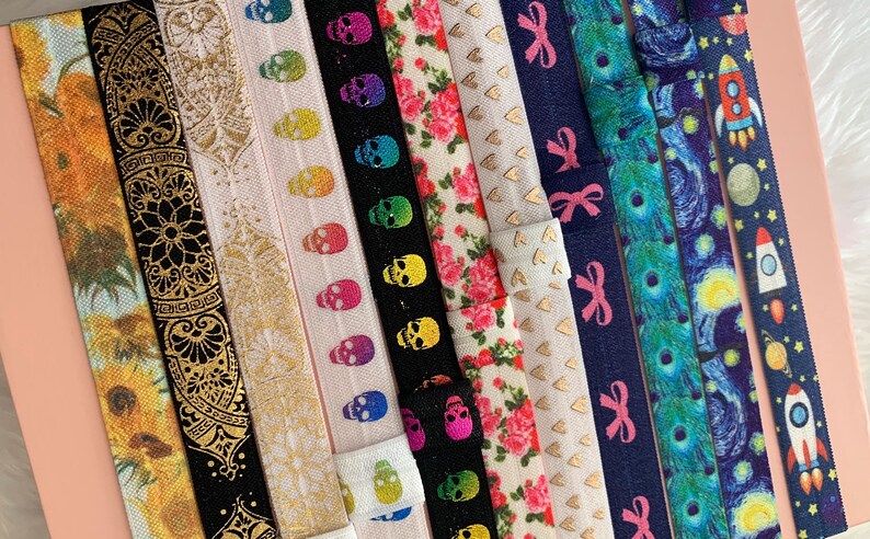 May include: A collection of colorful elastic hair ties with various patterns, including sunflowers, paisley, skulls, hearts, bows, and a starry night sky.