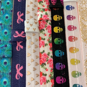 May include: Assortment of colorful elastic hair ties with various patterns, including a rocket ship, starry night, peacock feathers, bows, hearts, skulls, a mandala design, and sunflowers.