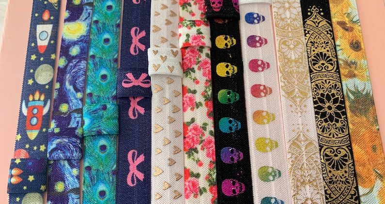 May include: Assortment of colorful elastic hair ties with various patterns, including a rocket ship, starry night, peacock feathers, bows, hearts, skulls, a mandala design, and sunflowers.