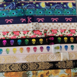 May include: A collection of colorful hair ties with various patterns, including stars, skulls, flowers, and geometric designs. The hair ties are arranged in a vertical stack on a pink surface.