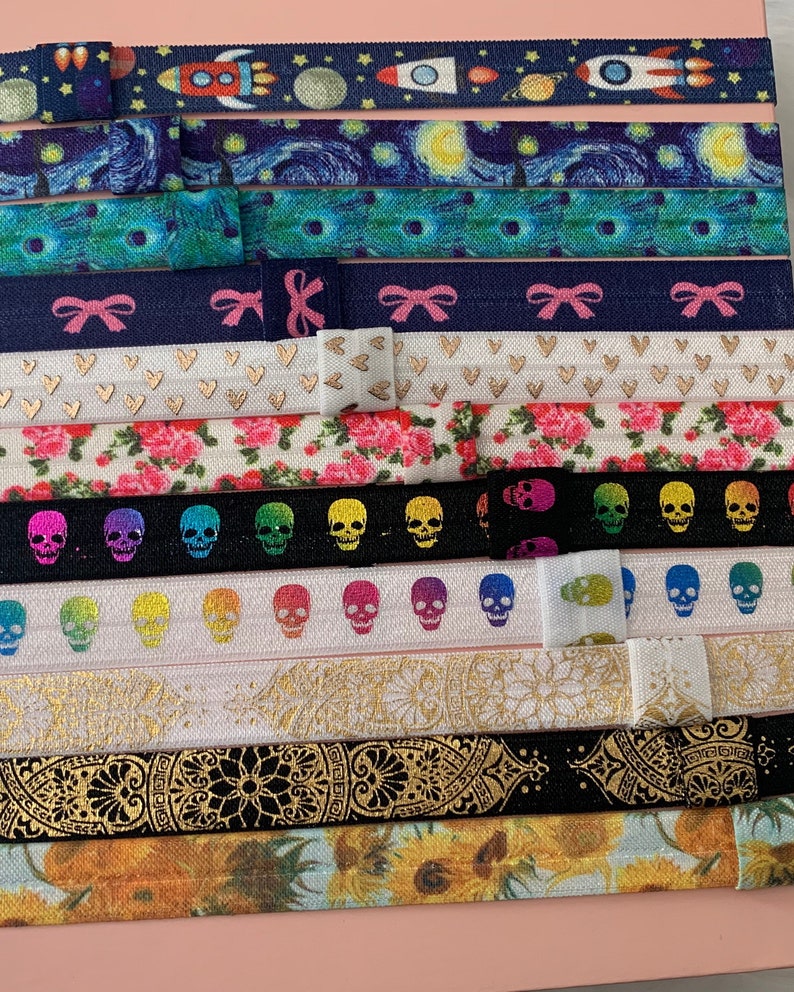May include: A collection of colorful hair ties with various patterns, including stars, skulls, flowers, and geometric designs. The hair ties are arranged in a vertical stack on a pink surface.