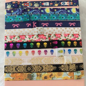 May include: A collection of 12 colorful hair ties with various patterns, including a starry night sky, a floral design, skulls, and a mandala. The hair ties are arranged in rows on a pink surface.