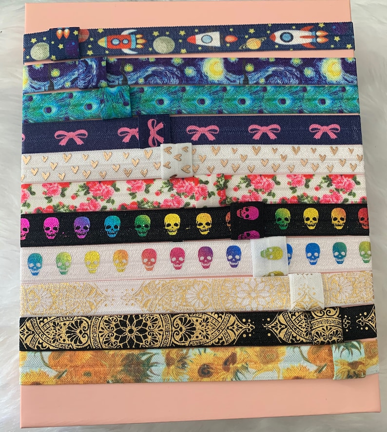 May include: A collection of 12 colorful hair ties with various patterns, including a starry night sky, a floral design, skulls, and a mandala. The hair ties are arranged in rows on a pink surface.