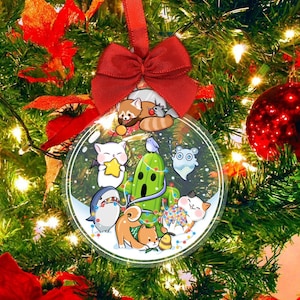 May include: A clear, round Christmas ornament with a red ribbon bow. The ornament features cartoon animals and a cactus decorated with lights. The background is a Christmas tree with lights and red ornaments.