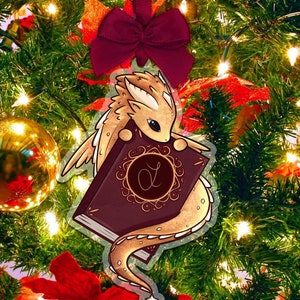 May include: A gold and brown dragon ornament holding a burgundy book with gold accents and a gold letter "D" on the cover. The ornament is hanging on a Christmas tree with red and gold decorations.