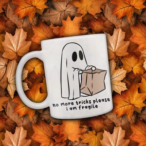 May include: White ceramic mug featuring a cartoon ghost holding a brown paper bag. The mug has the text "no more tricks please I am fragile". The background is a bed of autumn leaves in shades of orange and brown.