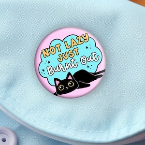 May include: A light blue backpack with a pink and white button that says "Not Lazy Just Burnt Out" with a black cat illustration.