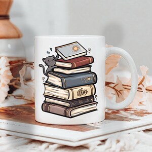 May include: White ceramic mug featuring a stack of books with a gray cat perched on top. The books are in various colors, with the text "tbr" visible on one. The top book has a sun design.