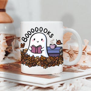 Op de afbeelding: Witte keramische mok met een wikkelontwerp met een cartoon spook dat een boek leest, omringd door herfstbladeren. De mok heeft het woord "BOOOOOOKS" erop gedrukt, en een kleine stapel boeken en een kopje koffie.