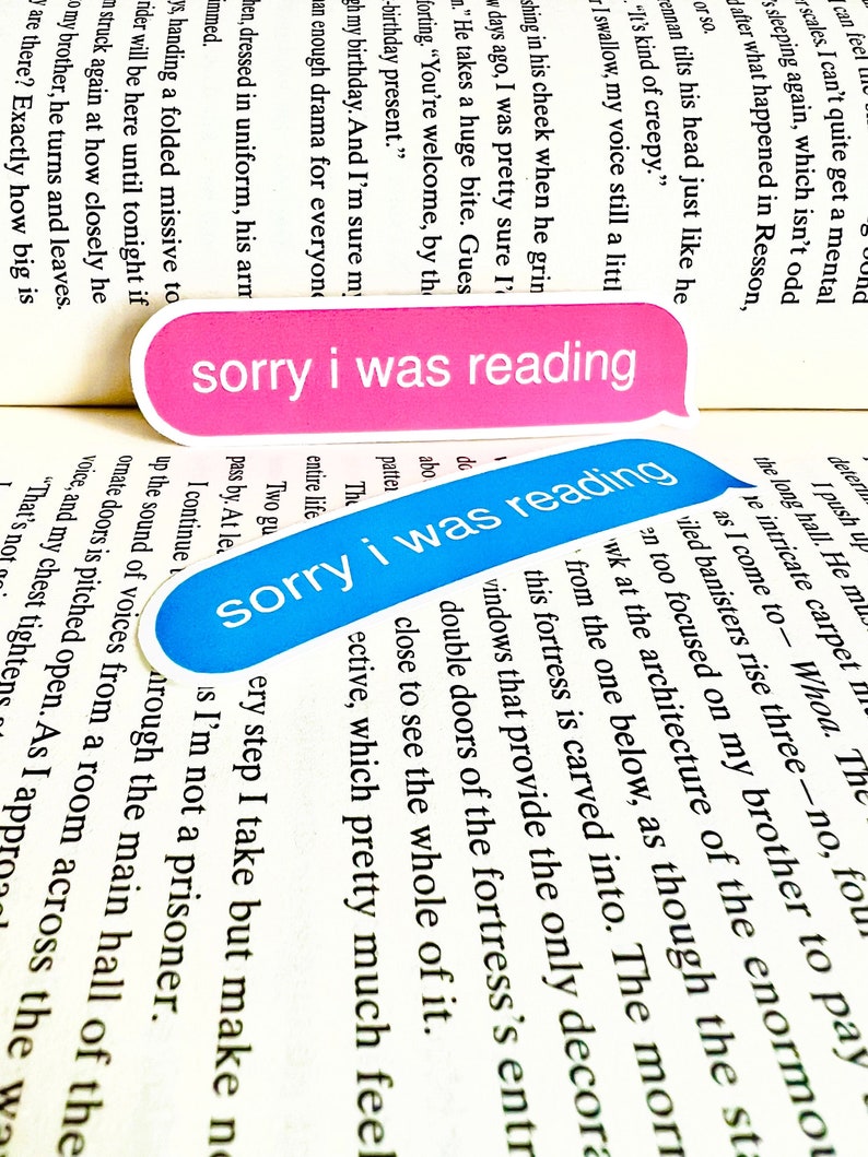 K&ouml;nnte beinhalten: Zwei blaue und rosafarbene Lesezeichen mit dem Text "sorry i was reading" darauf. Die Lesezeichen liegen auf einem ge&ouml;ffneten Buch.