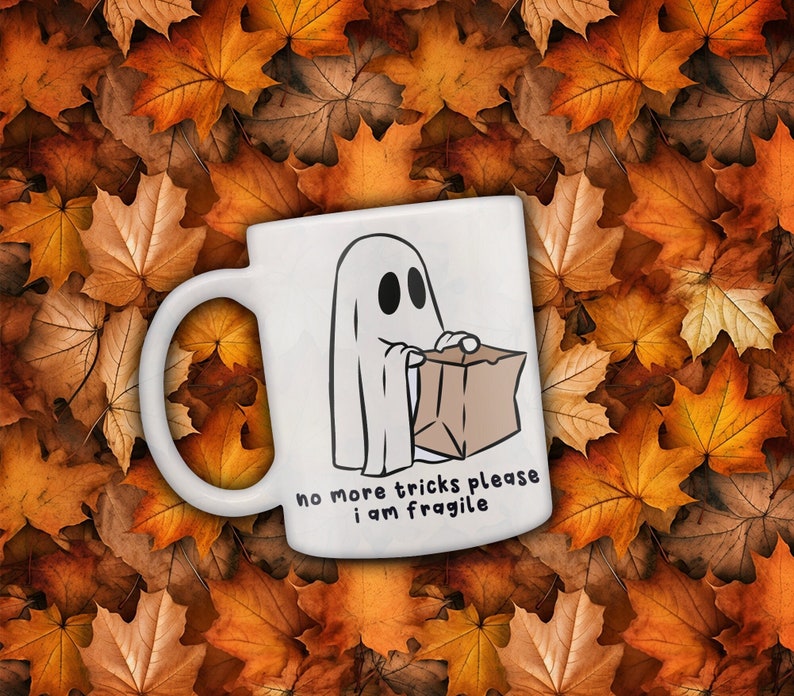 May include: White ceramic mug featuring a cartoon ghost holding a brown paper bag. The mug has the text "no more tricks please I am fragile". The background is a bed of autumn leaves in shades of orange and brown.