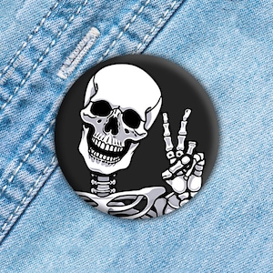 May include: A black and white button featuring a skeleton giving the peace sign. The skeleton is smiling and has a black background.