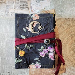 May include: A rectangular journal with a black fabric cover featuring a floral pattern in pink, yellow, and green. A silver-toned crescent moon and star charm is attached to the front. A dark red fabric ribbon wraps around the centre.