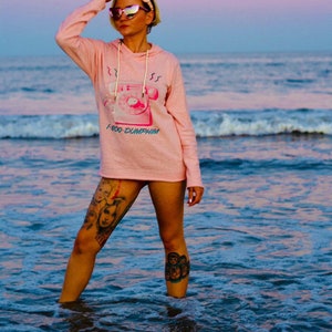 Puede incluir: Una mujer con una sudadera rosa con un gráfico de un teléfono rotatorio vintage y el texto "1-800-DUMPHIM". Está de pie en el océano con las piernas en el agua.