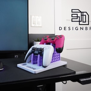 May include: A 3D printed purple stand holds two white and one pink PlayStation 5 controllers. The stand is labelled "3D DESIGNBROS".