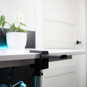 May include: A black desk clamp with a horizontal arm, mounted to a white desk. The clamp is used to hold a monitor or other device.