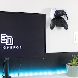 Op de afbeelding: Een aan de muur gemonteerde gamecontrollerhouder met een witte PlayStation 5-controller en een zwarte Xbox-controller. De houder is aan een witte muur bevestigd, met een lichtschakelaar en een kleine potplant.