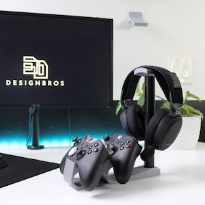 Puede incluir: Un soporte blanco con dos mandos de videojuegos negros y unos auriculares negros. El soporte está sobre un escritorio blanco con una alfombrilla de ratón negra y un ratón negro. El fondo es una pared blanca con un monitor negro que muestra el texto "DESIGNBROS".