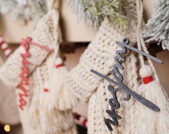 May include: Two white knitted stockings with tassels hanging on a fireplace mantel. One stocking has a red and white candy cane hanging from it. The other stocking has a black wooden sign with the word "Merry" written on it.