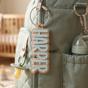 May include: A sage green quilted diaper bag with a matching pacifier and bottle. A custom name tag that says "HARPER" in light blue letters hangs from the bag. The bag has multiple pockets and a shoulder strap.