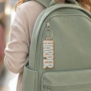 May include: A rectangular keychain featuring the name HARPER in raised light blue letters on a tan background, attached to a silver metal split ring. The keychain is clipped to the zipper pull of a sage green backpack with multiple compartments.
