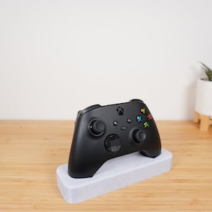 May include: A black Xbox controller is sitting on a white charging stand. The controller has a black and white design with a green, yellow, red, and blue button.