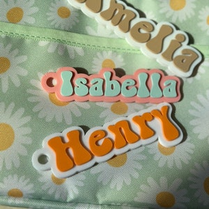 May include: Three personalized name tags with a retro font style. The names are Amelia, Isabella, and Henry. The tags are made of white plastic with colorful letters.