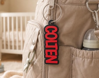 May include: A beige quilted backpack featuring a black and red rectangular keychain with the name COLTEN printed in bold red capital letters. The keychain is attached to the silver zipper pull of the backpack's front pocket.