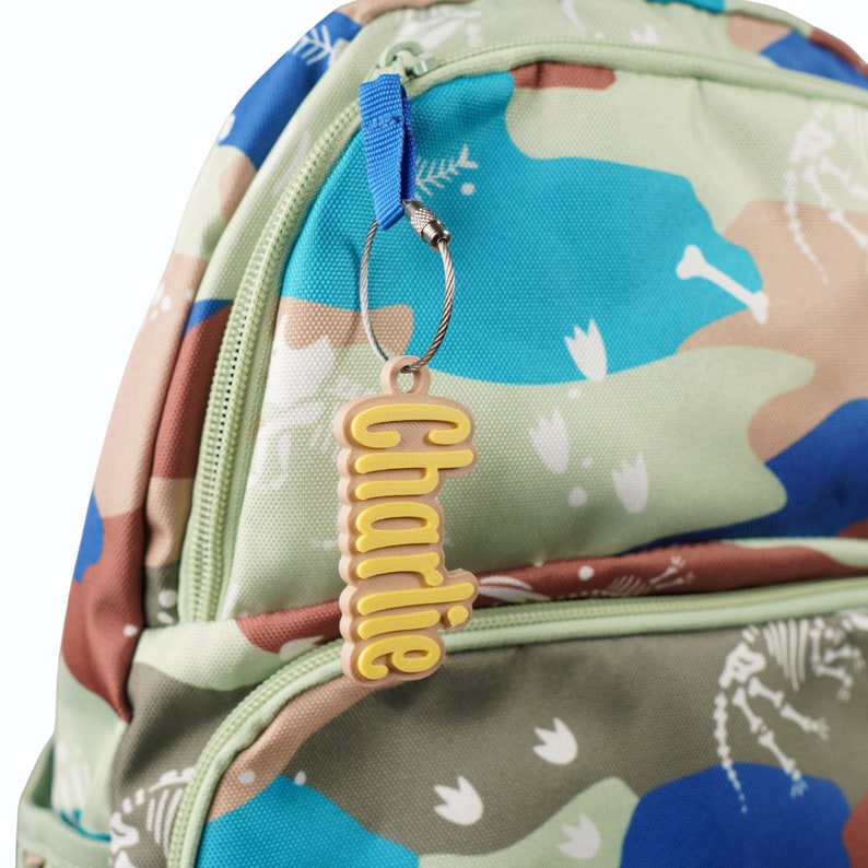 May include: A green, blue and brown camouflage backpack with a yellow name tag that says "Charlie" attached to it.