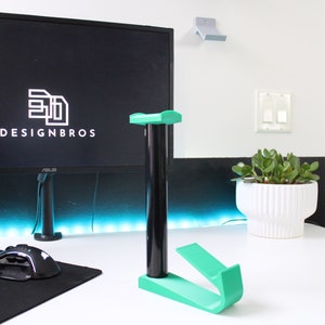 May include: A green plastic headset stand with a black base. The stand is on a white desk with a black keyboard, a black mouse, and a black computer monitor with the text "3D DESIGNBROS" on the screen.