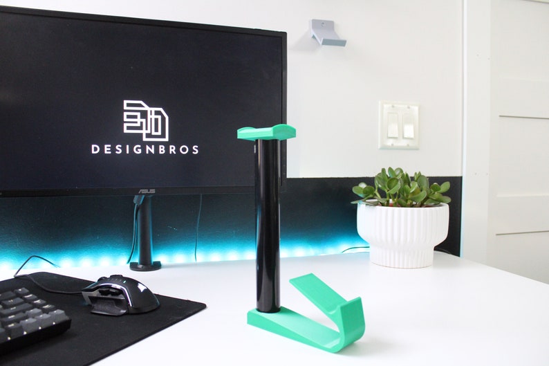 May include: A green plastic headset stand with a black base. The stand is on a white desk with a black keyboard, a black mouse, and a black computer monitor with the text "3D DESIGNBROS" on the screen.